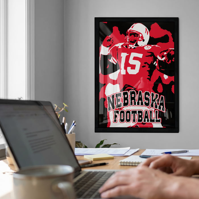 a framed poster of a football player, with the text "NEBRASKA FOOTBALL" prominently displayed. In the foreground, there is a laptop computer and a coffee cup on a desk, suggesting a workspace or home office setting.