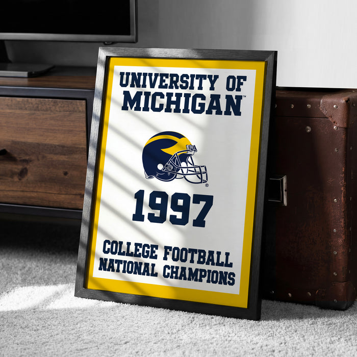 A framed poster with the text "UNIVERSITY OF MICHIGAN 1997 COLLEGE FOOTBALL NATIONAL CHAMPIONS" and a football helmet graphic, placed on a carpeted floor next to a wooden chest.