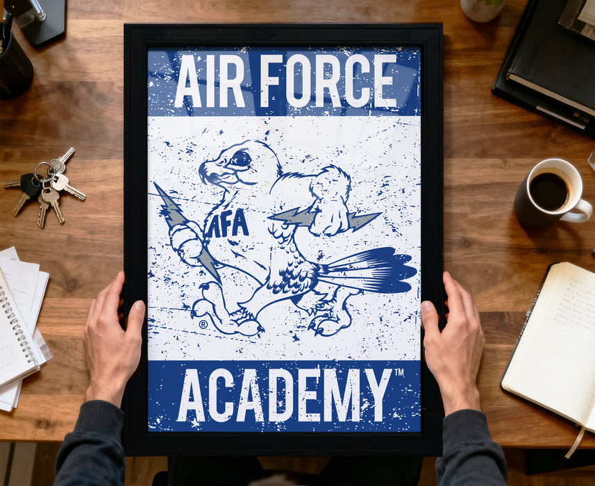 A framed poster of an Air Force Academy logo is being held by a person's hands, with a wooden desk and various items in the background.