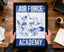 A framed poster of an Air Force Academy logo is being held by a person's hands, with a wooden desk and various items in the background.