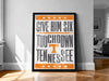 A framed poster with the text "GIVE HIM SIX TOUCHDOWN TENNESSEE" on it, placed on a wooden floor against a white wall.