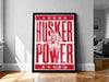 A framed poster with the text "HUSKER POWER" and a cartoon character, placed on a wooden floor against a white wall.
