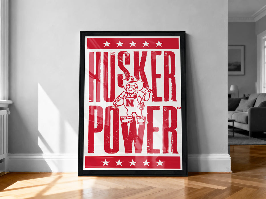 A framed poster with the text "HUSKER POWER" and a cartoon character, placed on a wooden floor against a white wall.