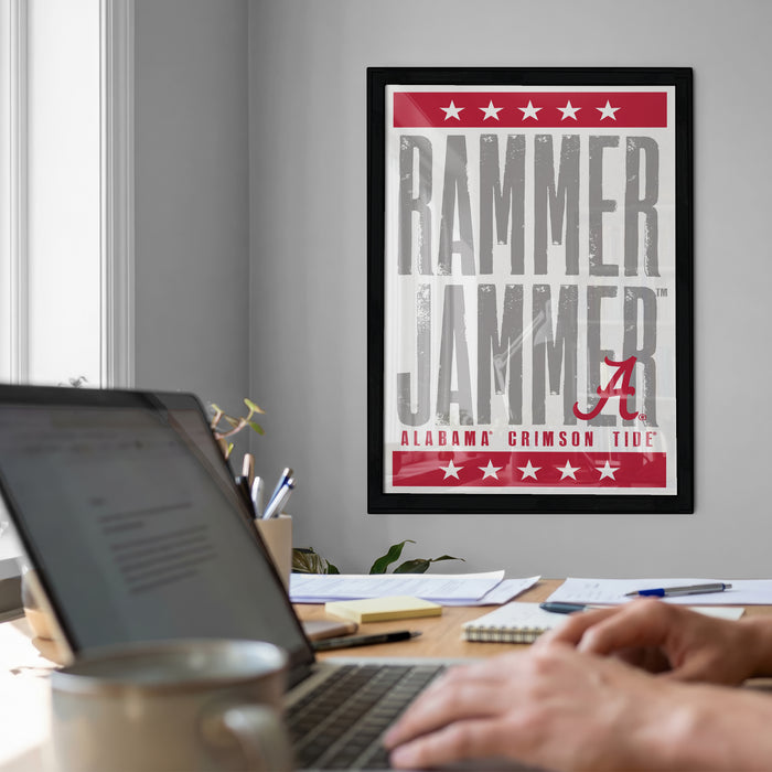 a framed poster with the text "RAMMER JAMMER" and "ALABAMA GRIISON TIDE" on it, hanging on a wall above a desk with a laptop and other items on it.