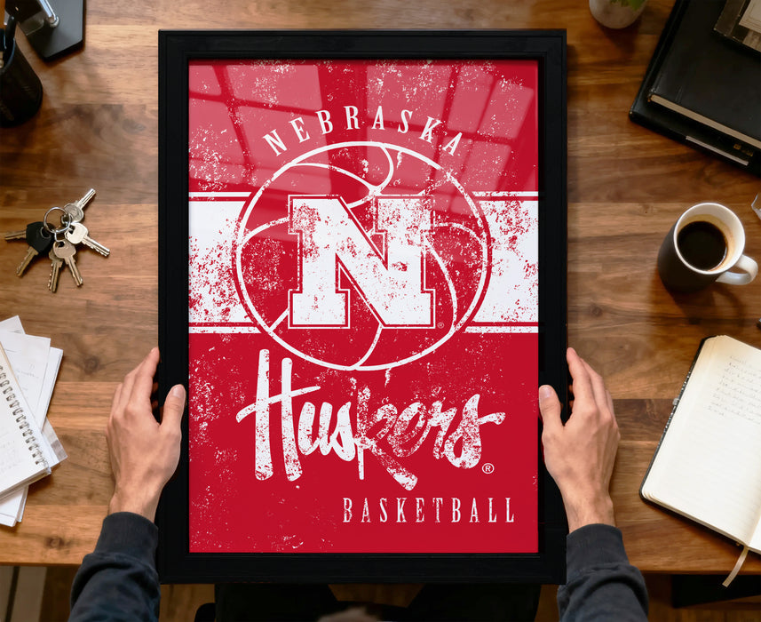 A person is holding up a framed poster of the Nebraska Huskers basketball team, which features the team's logo and name.