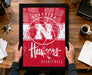 A person is holding up a framed poster of the Nebraska Huskers basketball team, which features the team's logo and name.