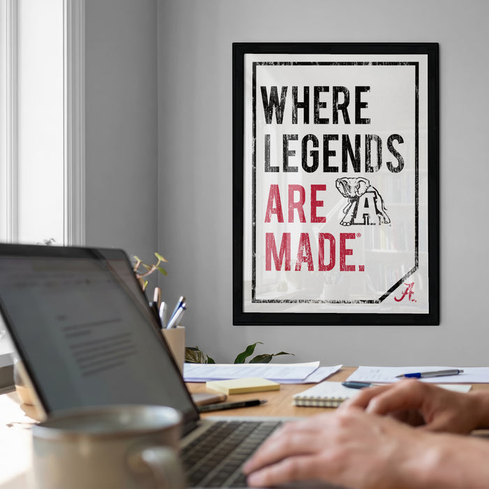 a framed motivational poster with the text "WHERE LEGENDS ARE MADE." in bold, black and red lettering, along with an illustration of an elephant. The poster is displayed on a wall above a desk with a laptop, a coffee mug, and some papers.