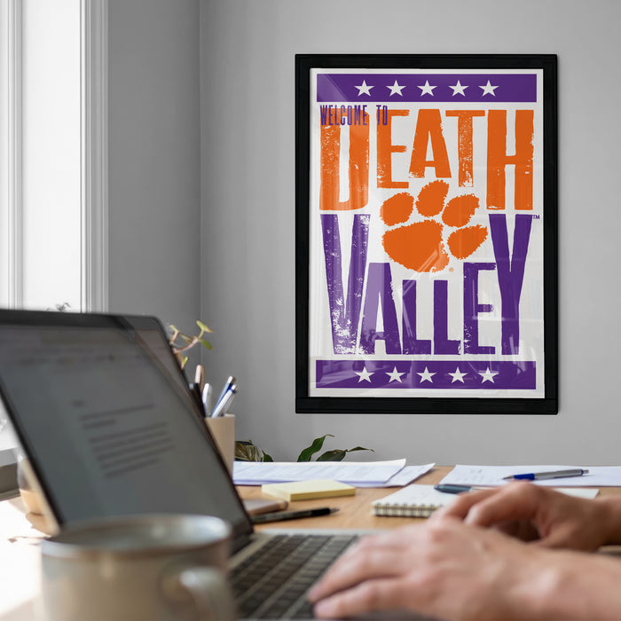 a person working on a laptop computer, with a framed poster of "Welcome to Death Valley" hanging on the wall behind them.