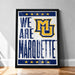 A framed poster with the text "WE ARE MARQUETTE" and the Marquette University logo, placed on a wooden floor against a white wall.