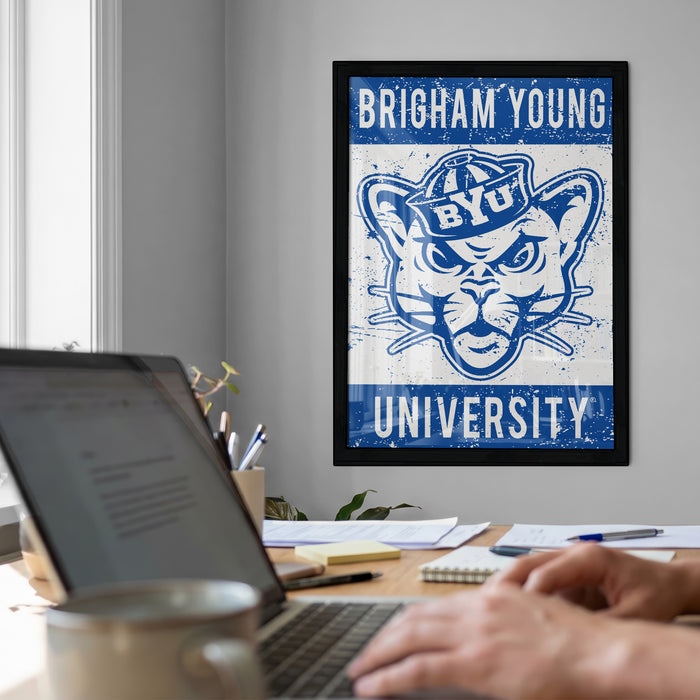 A framed poster of the University of Utah's mascot is displayed on the wall above a desk with a laptop, a coffee mug, and some papers.