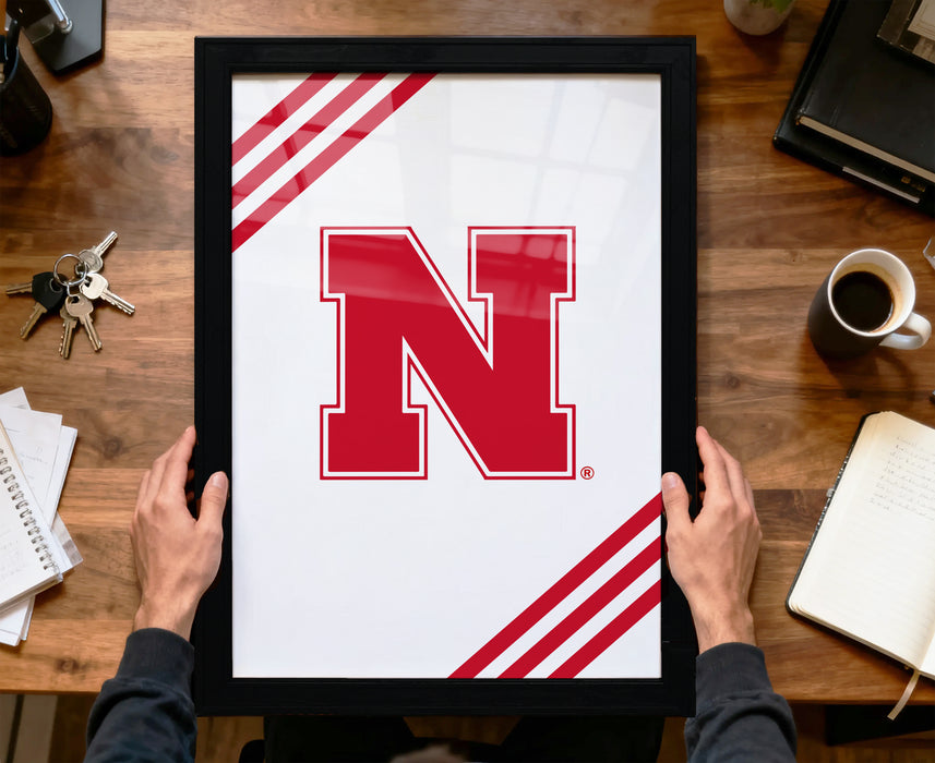 A person is holding a framed picture with a large red letter "N" on it, which appears to be a logo or emblem. The picture is placed on a wooden desk, along with various items such as a cup, a notebook, and a set of keys.