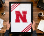 A person is holding a framed picture with a large red letter "N" on it, which appears to be a logo or emblem. The picture is placed on a wooden desk, along with various items such as a cup, a notebook, and a set of keys.
