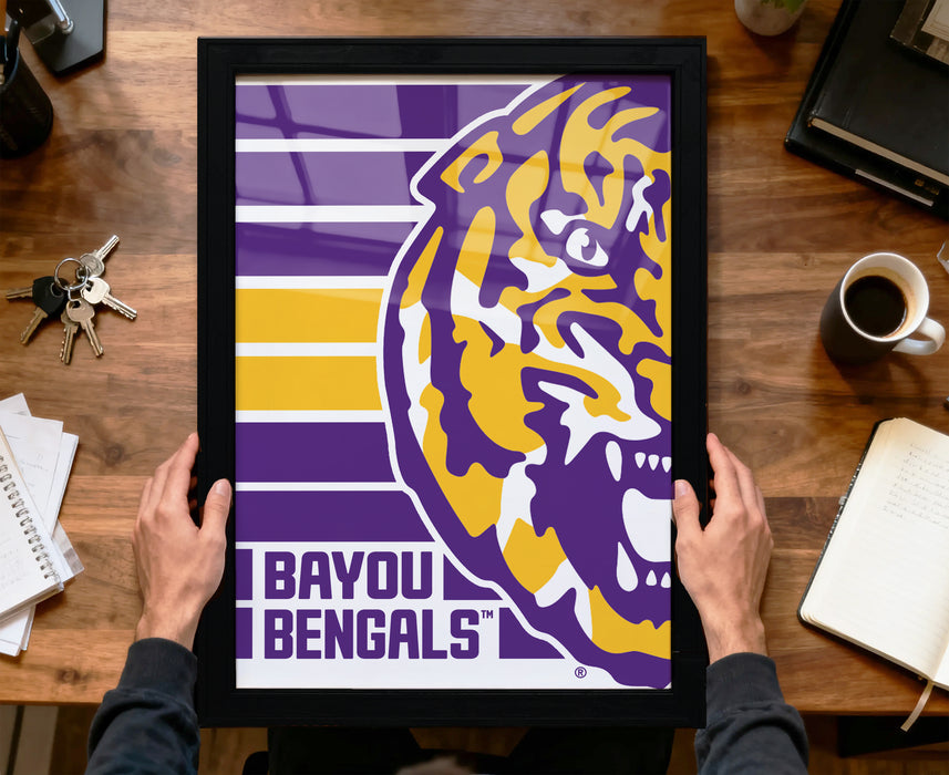 A framed poster of the LSU (Louisiana State University) logo is being held by a person's hands, with a wooden desk and various items such as keys, a cup, and a notebook in the background.