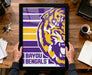 A framed poster of the LSU (Louisiana State University) logo is being held by a person's hands, with a wooden desk and various items such as keys, a cup, and a notebook in the background.