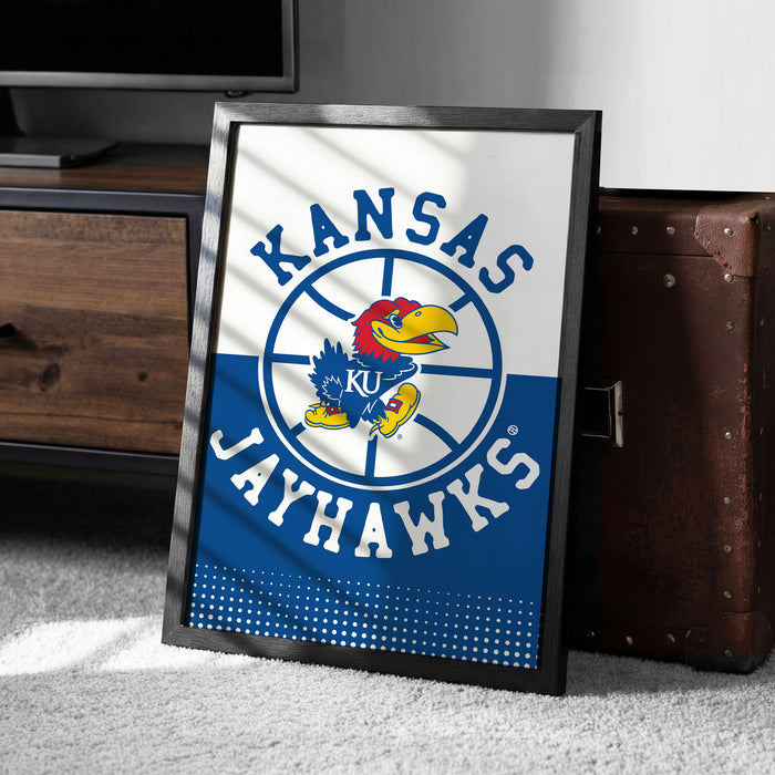 A framed poster of the Kansas Jayhawks basketball team, featuring the team's logo and name, is displayed on a carpeted floor next to a wooden chest.