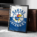 A framed poster of the Kansas Jayhawks basketball team, featuring the team's logo and name, is displayed on a carpeted floor next to a wooden chest.