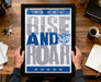 A framed poster with the text "RISE AND ROAR" and a cat logo is being held by a person's hands.