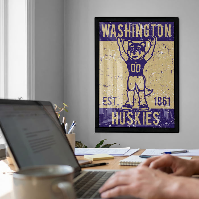 a framed poster of a mascot, a dog, with the text "WASHINGTON EST. 1861 HUSKIES" on it, hanging on a wall above a desk with a laptop and other items.