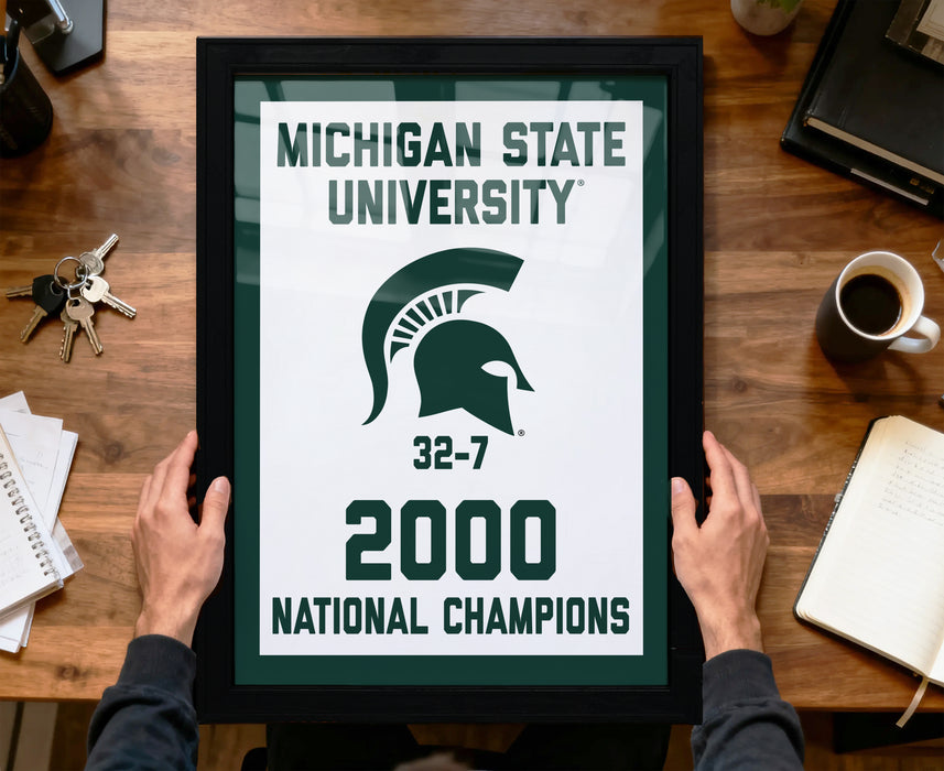 A framed poster with the text "MICHIGAN STATE UNIVERSITY" and "32-7 2000 NATIONAL CHAMPIONS" is being held by a person's hands.