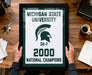 A framed poster with the text "MICHIGAN STATE UNIVERSITY" and "32-7 2000 NATIONAL CHAMPIONS" is being held by a person's hands.