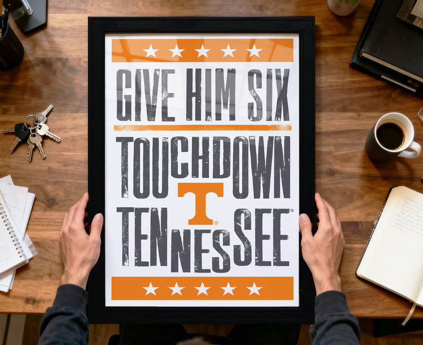 A framed poster with the text "GIVE HIM SIX TOUCHDOWN TENNESSEE" is being held by a person's hands.