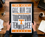 A framed poster with the text "GIVE HIM SIX TOUCHDOWN TENNESSEE" is being held by a person's hands.
