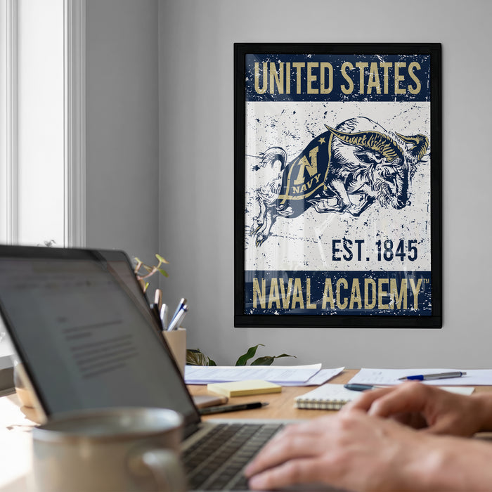 a framed poster of a naval emblem hanging on a wall, with a laptop and a person's hands visible in the foreground.