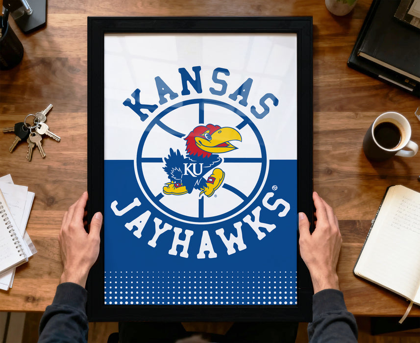 A framed poster of the Kansas Jayhawks basketball team is being held by a person's hands, with a wooden desk and various items such as keys, a coffee cup, and a notebook in the background.