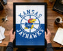 A framed poster of the Kansas Jayhawks basketball team is being held by a person's hands, with a wooden desk and various items such as keys, a coffee cup, and a notebook in the background.
