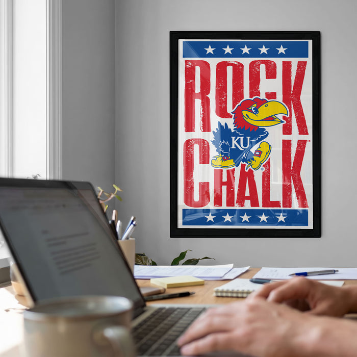a framed poster of a bird, likely a bird of prey, with the text "ROCK CHALK" prominently displayed. In the foreground, there is a laptop computer and a coffee mug on a desk, suggesting a workspace or home office setting.