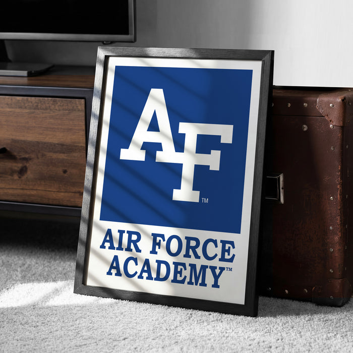 a framed poster with the Air Force Academy logo, placed on a carpeted floor next to a wooden chest.
