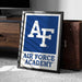 a framed poster with the Air Force Academy logo, placed on a carpeted floor next to a wooden chest.