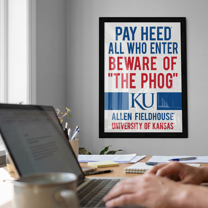 a framed poster on a wall with the text "PAY HEED ALL WHO ENTER BEWARE OF 'THE PHOG' KU Allen Fieldhouse UNIVERSITY OF KANSAS" displayed prominently. In the foreground, there is a laptop and a coffee cup on a desk, suggesting a workspace or office setting.