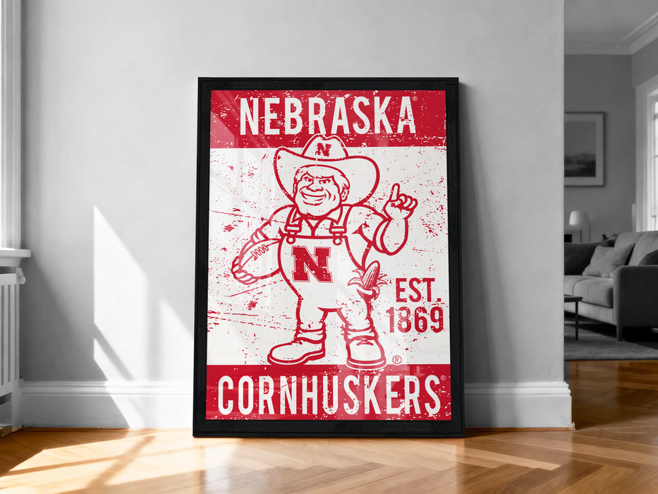 A framed poster of a Cornhuskers mascot, a red and white cartoon character, is displayed on a wall in a room.