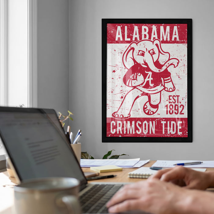 a framed poster of an elephant with the text "ALABAMA" and "CRIMSON TIDE" on it, hanging on a wall above a desk with a laptop and other items.