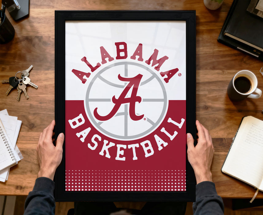 A framed poster of the Alabama basketball team logo is being held by a person's hands, with a desk and various items such as keys, a cup, and a notebook visible in the background.