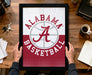 A framed poster of the Alabama basketball team logo is being held by a person's hands, with a desk and various items such as keys, a cup, and a notebook visible in the background.