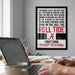 a framed poster with text on it, hanging on a wall above a desk with a laptop and other items on it.