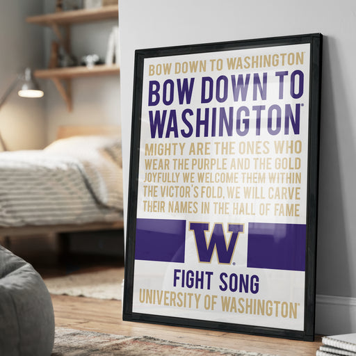 The image is a framed poster with text that reads "BOW DOWN TO WASHINGTON BOW DOWN TO WASHINGTON MIGHTY ARE THE ONES WHO WEAR THE PURPLE AND THE GOLD JOYFULLY WE WELCOME THEM WITHIN THE VICTOR'S FOLD. WE WILL CARVE THEIR NAMES IN THE HALL OF FAME". The poster is displayed on a wall in a room, with a bed and a bookshelf visible in the background.