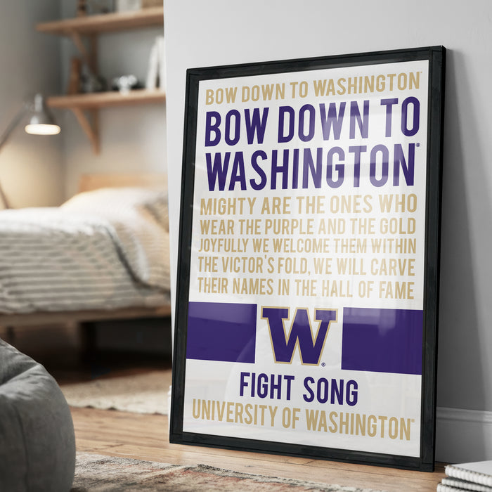 The image is a framed poster with text that reads "BOW DOWN TO WASHINGTON BOW DOWN TO WASHINGTON MIGHTY ARE THE ONES WHO WEAR THE PURPLE AND THE GOLD JOYFULLY WE WELCOME THEM WITHIN THE VICTOR'S FOLD. WE WILL CARVE THEIR NAMES IN THE HALL OF FAME". The poster is displayed on a wall in a room, with a bed and a bookshelf visible in the background.