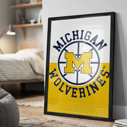 A framed poster of the Michigan Wolverines basketball team logo is displayed on a wall in a bedroom.