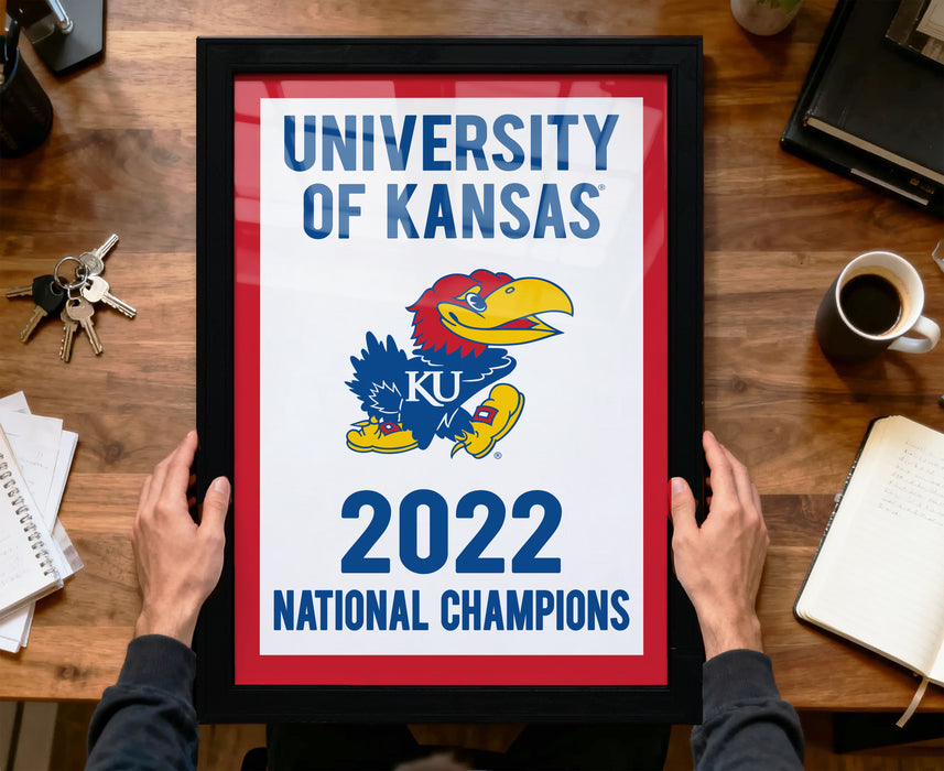 A framed poster with the text "UNIVERSITY OF KANSAS" and "2022 NATIONAL CHAMPIONS" is being held by a person's hands.