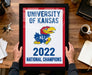 A framed poster with the text "UNIVERSITY OF KANSAS" and "2022 NATIONAL CHAMPIONS" is being held by a person's hands.