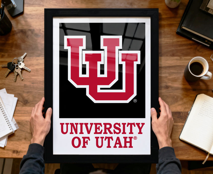 A person is holding up a framed poster of the University of Utah logo on a wooden desk.