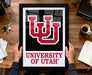 A person is holding up a framed poster of the University of Utah logo on a wooden desk.