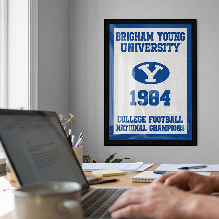 A framed poster of Brigham Young University from 1984 hangs on a wall, with a laptop and coffee cup on a desk in the foreground.
