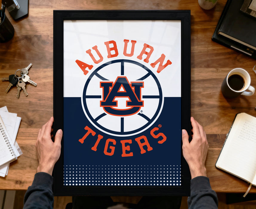 A framed poster of the Auburn Tigers logo is being held by a person's hands, with a wooden desk and various items such as keys, a cup, and a notebook in the background.