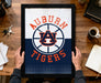 A framed poster of the Auburn Tigers logo is being held by a person's hands, with a wooden desk and various items such as keys, a cup, and a notebook in the background.