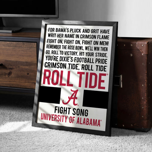 a framed poster with text that reads "For Bama's Pluck and Grit Have WRIT HER NAME IN CRIMSON FLAME FIGHT ON, FIGHT ON, FIGHT ON MEN! REMEMBER THE ROSE BOWL, WE'LL WIN THEN GO, ROLL TO VICTORY, HIT YOUR STRIDE, YOU'RE DIXIE'S FOOTBALL PRIDE CRIMSON TIDE, ROLL TIDE". The poster is placed on a carpeted floor next to a wooden chest.