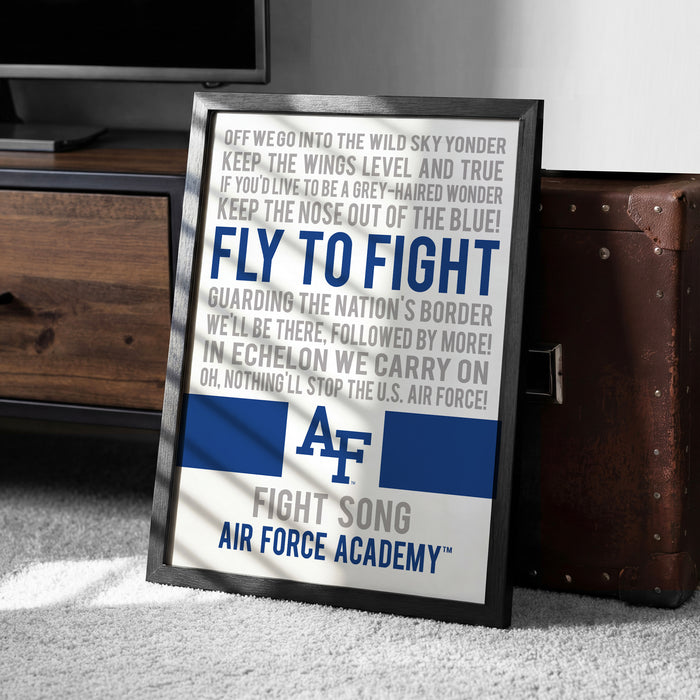 a framed poster with the text "AF FIGHT SONG AIR FORCE ACADEMY" on it, placed on a carpeted floor next to a wooden chest.